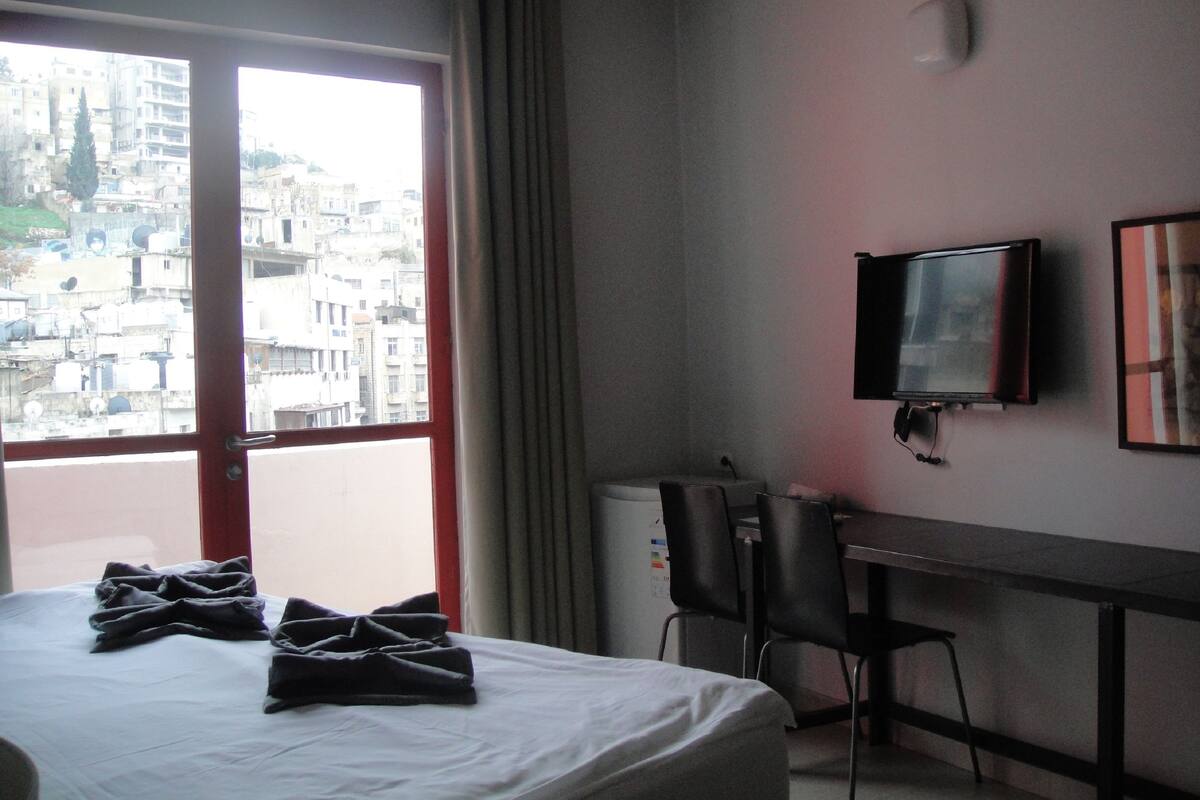 A hotel room with a bed, a desk, a chair, a television, and a view of a cityscape through a large window.
