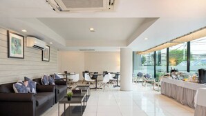 Un moderno hall de hotel con un sofá de cuero oscuro, sillas blancas, una estación de café y grandes ventanales con vistas a la ciudad.