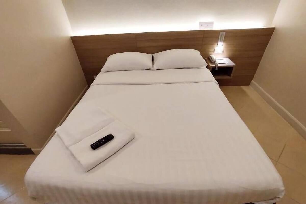 Un lit simple avec une literie blanche, une télécommande posée sur le lit et une table de chevet avec une lampe.
