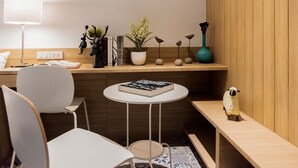 Una habitación moderna y luminosa con un escritorio de madera, una silla blanca, una pequeña mesa redonda y una estantería con adornos, entre ellos una planta en maceta y figuritas.