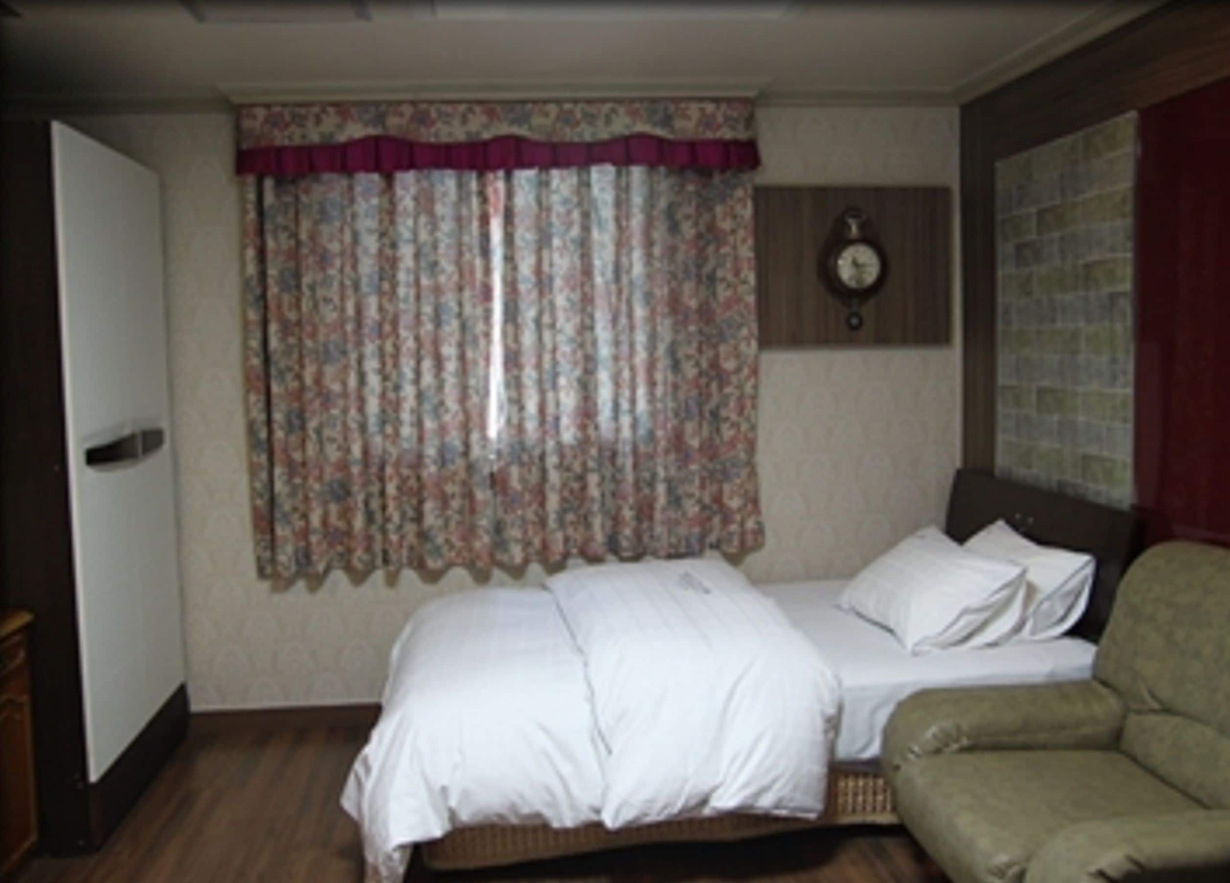 A bedroom with a bed, a chair, a clock, and a window with curtains.