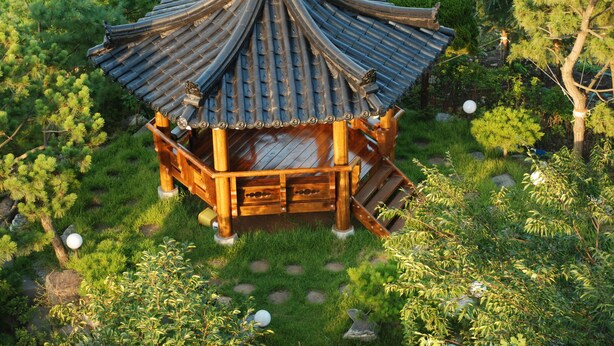 Um gazebo em estilo japonês tradicional, com telhado de telhas, rodeado por vegetação exuberante e pedras.
