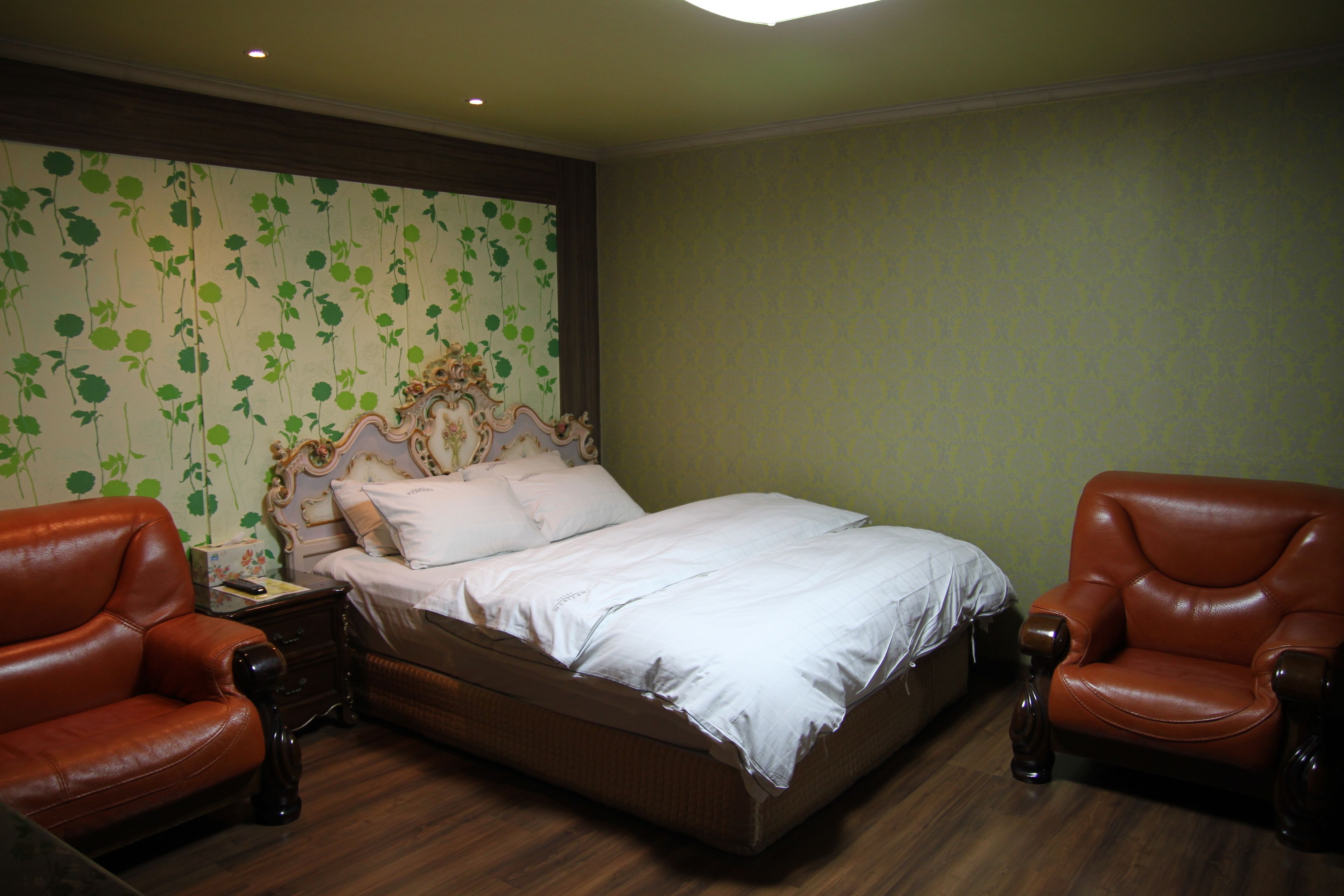 Ein Schlafzimmer mit einem Bett, zwei braunen Ledersesseln, einem Nachttisch und einer Wand mit floralem Wandpapier.