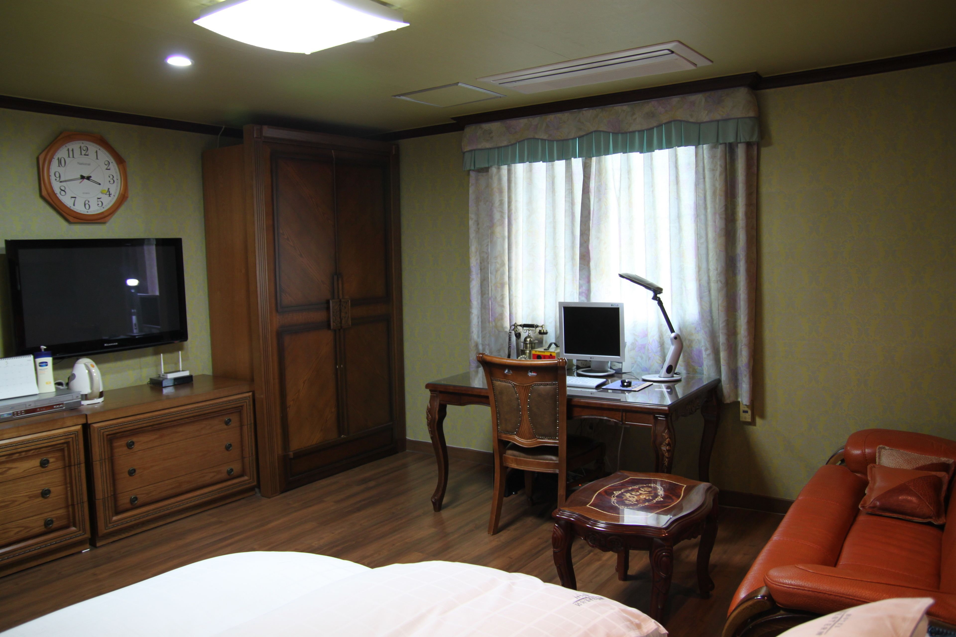 A hotel room with a bed, a desk with a computer, a television, a chair, a sofa, and a clock on the wall.