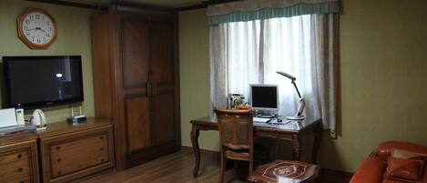 A hotel room with a bed, a desk with a computer, a television, a chair, a sofa, and a clock on the wall.