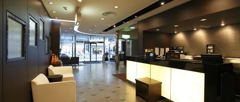 A modern hotel lobby with a reception desk, seating area, and a view of the street.