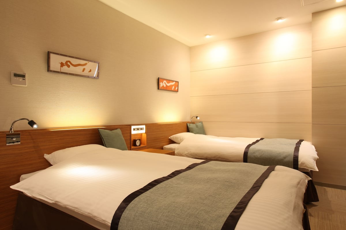 A hotel room with two beds, a nightstand, a lamp, and a wall-mounted clock.