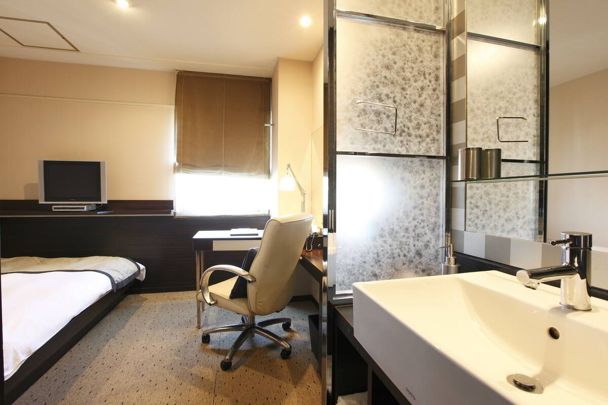 A hotel room with a bed, desk, chair, TV, and bathroom with a sink and mirror.