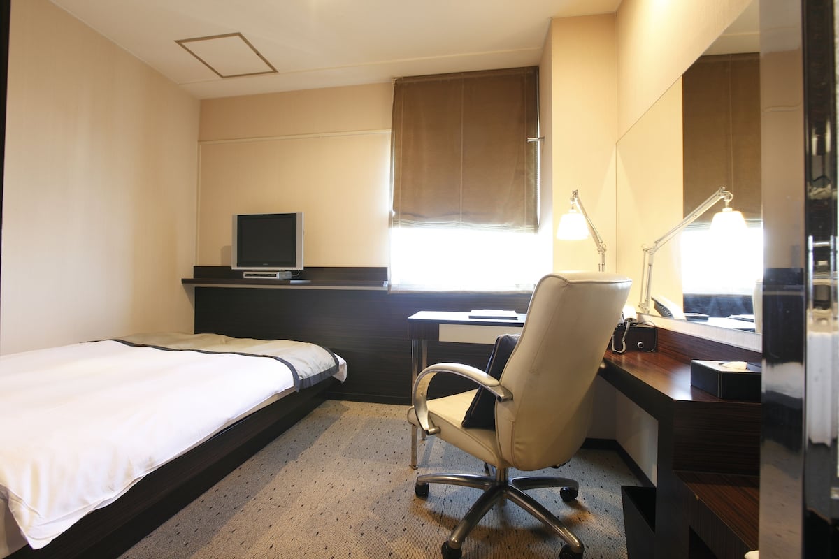 A hotel room with a bed, desk, chair, TV, and a window with blinds.