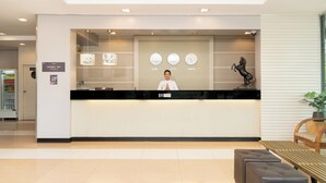 Een hotel receptiebalie met een receptionist, een klok die de tijd in drie steden weergeeft, en een beeld van een paard.