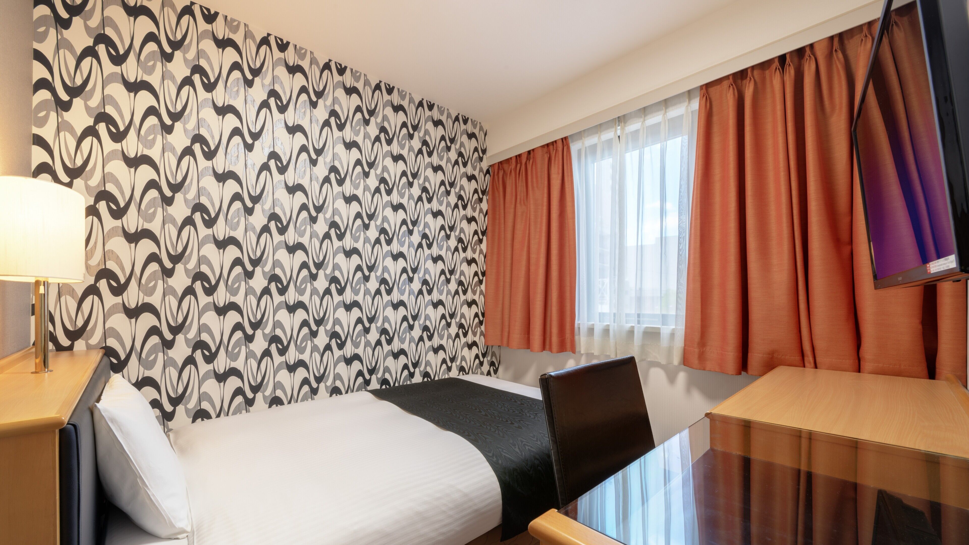 A hotel room with a patterned wallpaper, a bed, a desk with a laptop, a TV, and a window with curtains.