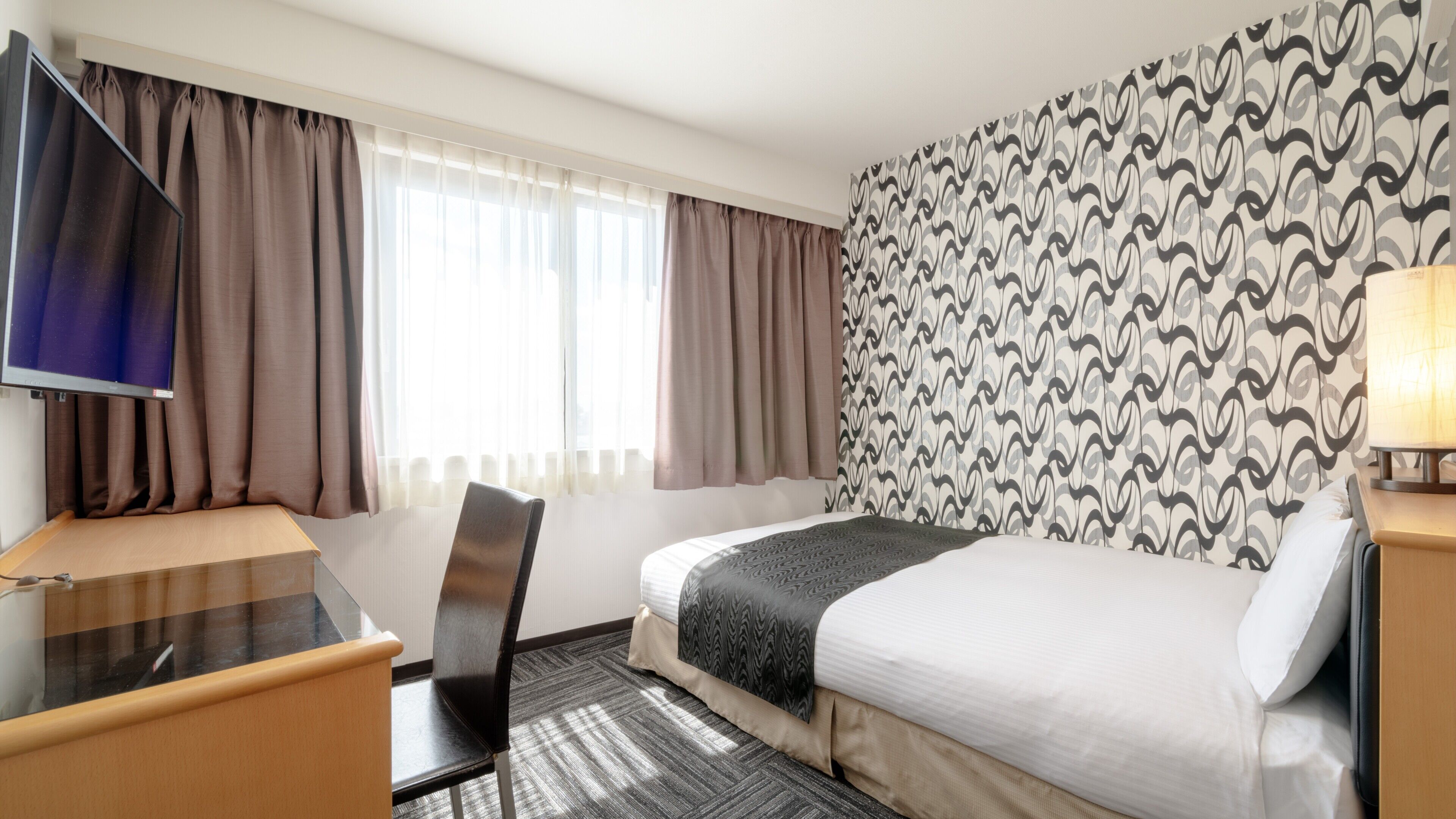 A hotel room with a bed, a desk, a chair, a TV, and a patterned wall.