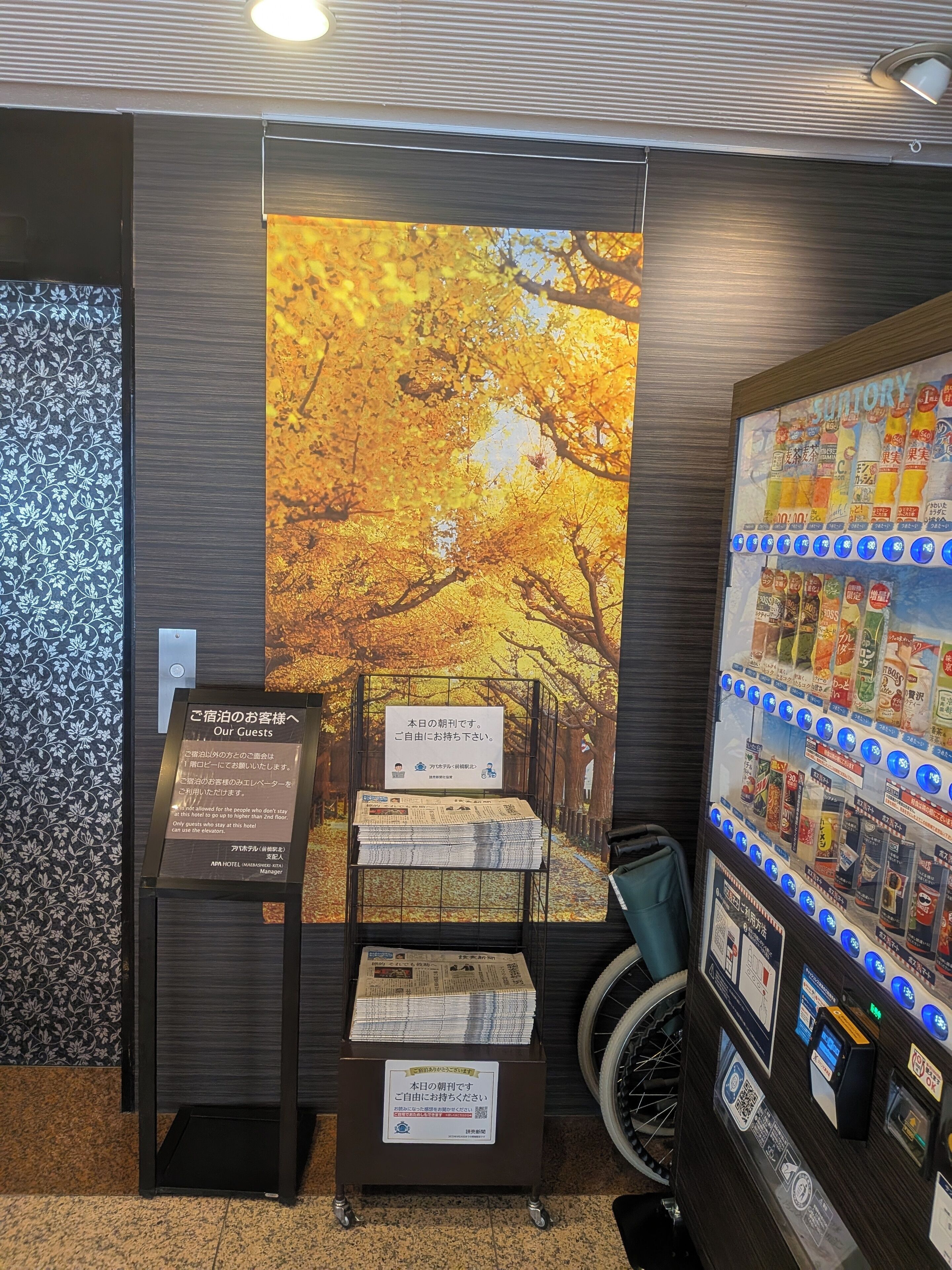 A hotel reception area with a large autumn-themed poster, a display of newspapers, and a vending machine.