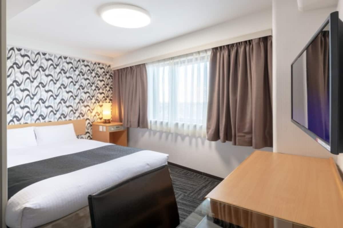 Une chambre d’hôtel avec un lit, un bureau, une télévision, une lampe et une fenêtre avec des rideaux.