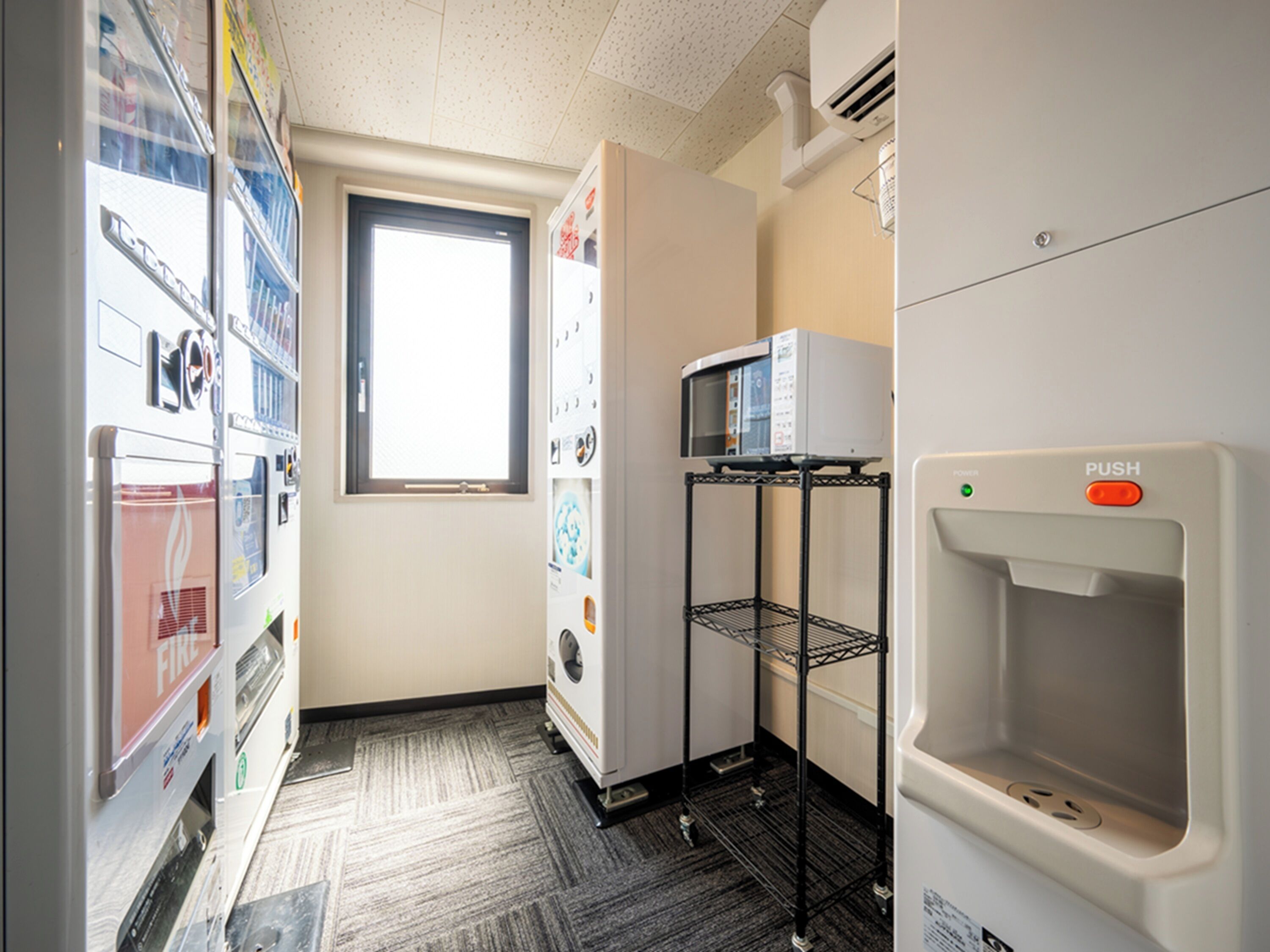 Quarto de hotel com cozinha compacta, micro-ondas numa prateleira, dispensador de água e janela com luz natural.