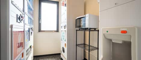A hotel room with a kitchenette, a microwave on a shelf, a water dispenser, and a window with natural light.