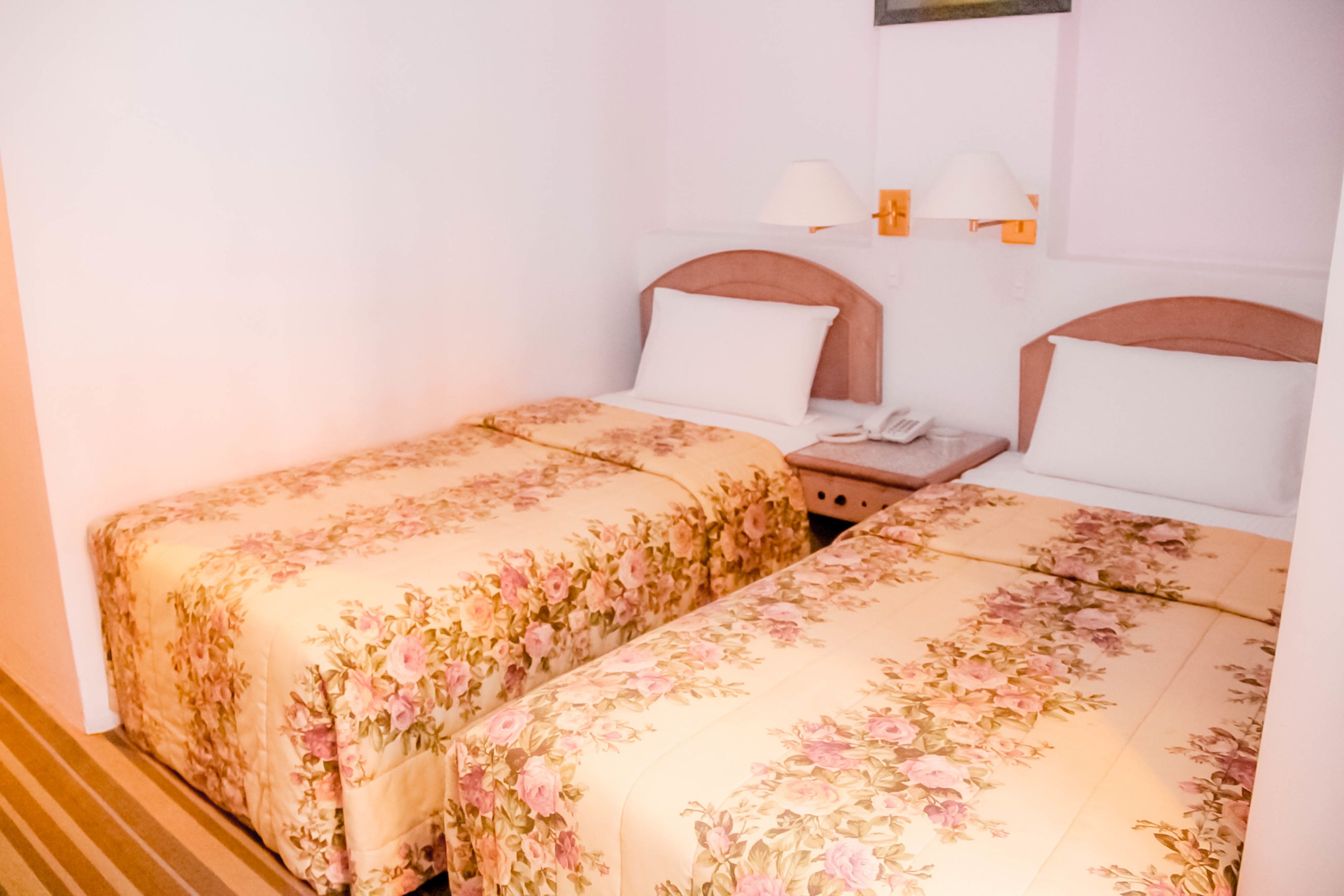 A hotel room with two beds, each with floral bedding, a bedside table with a lamp, and a wall-mounted telephone.