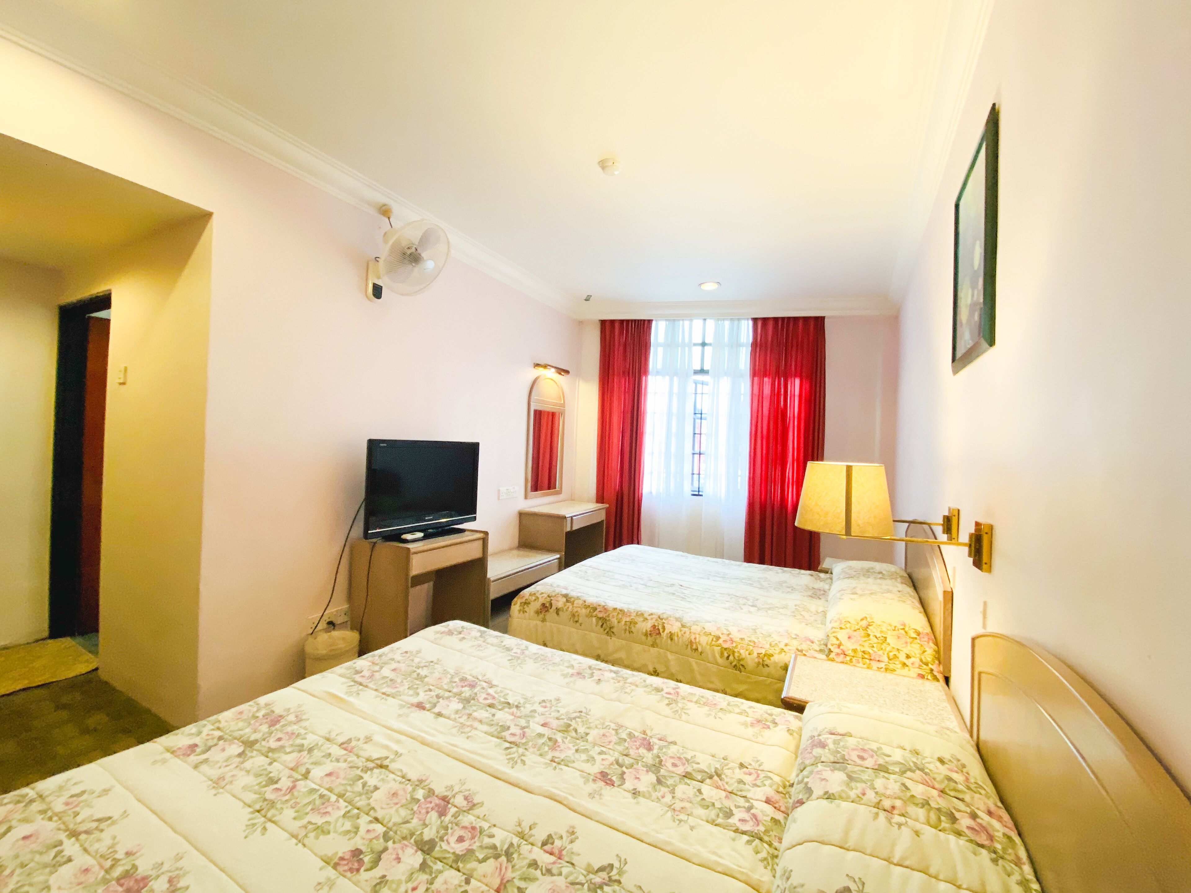 Une chambre d’hôtel avec deux lits, un bureau avec une télévision, une lampe et une fenêtre avec des rideaux rouges.