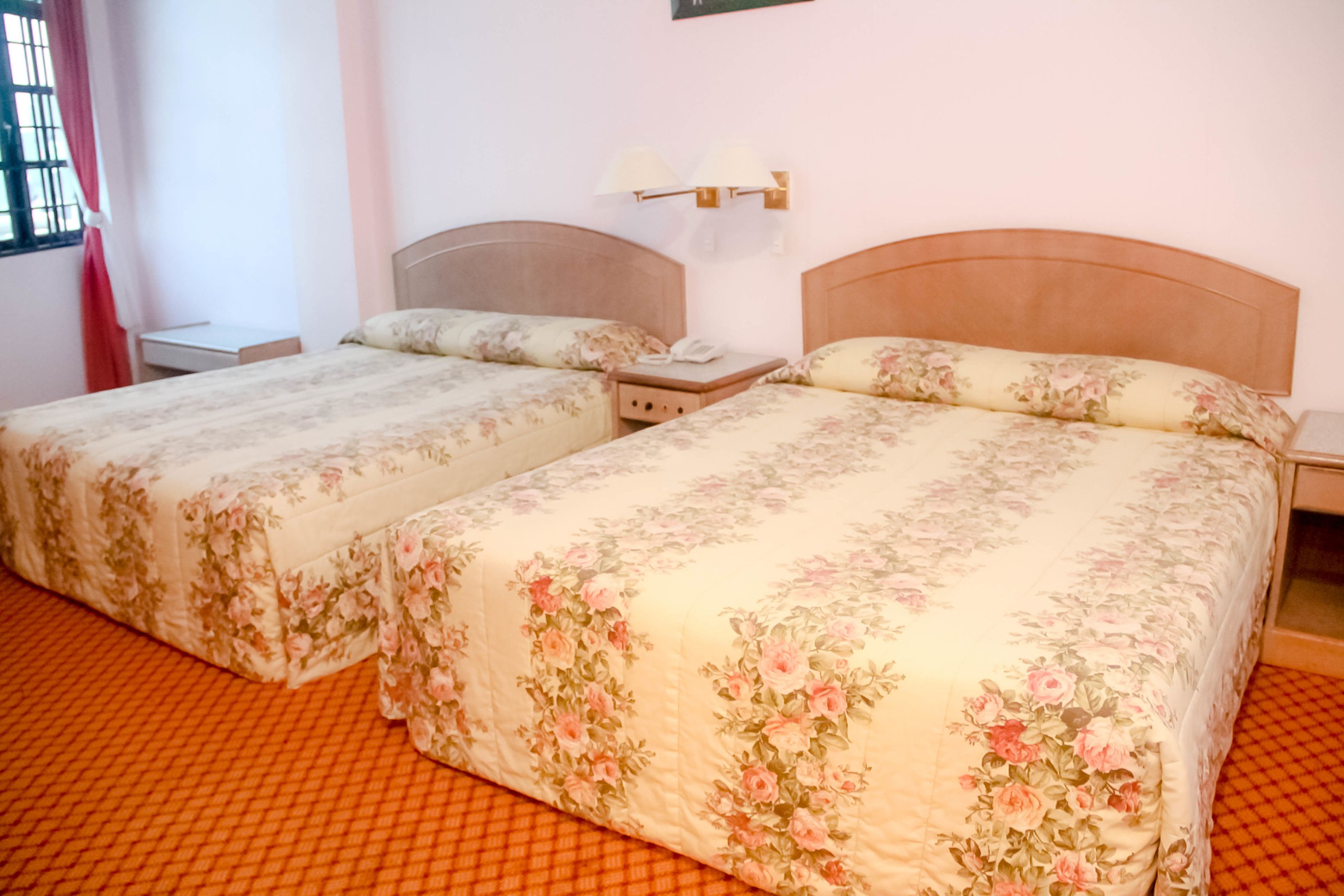 Two single beds with floral bedding, a nightstand with a lamp, and a window with a curtain.