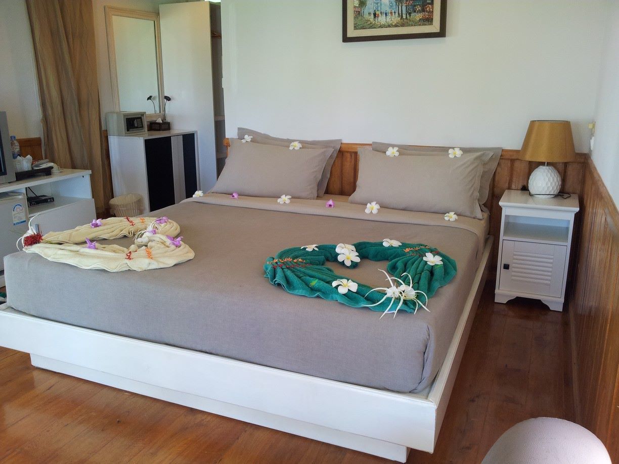 A neatly made bed with a grey bedspread, two pillows, and a green floral headboard. There is a bedside table with a lamp and a small decorative item.