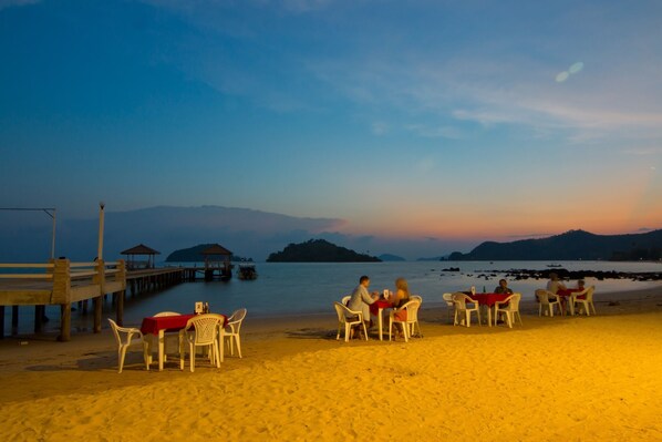 A beachside dining area with tables and chairs, overlooking a calm sea and distant islands.