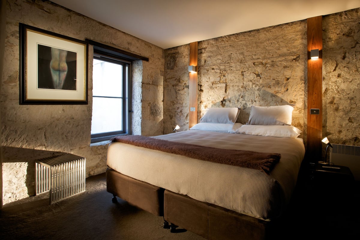 A bedroom with a stone wall, a framed picture, a window, a bed with a brown headboard, and a bedside table.