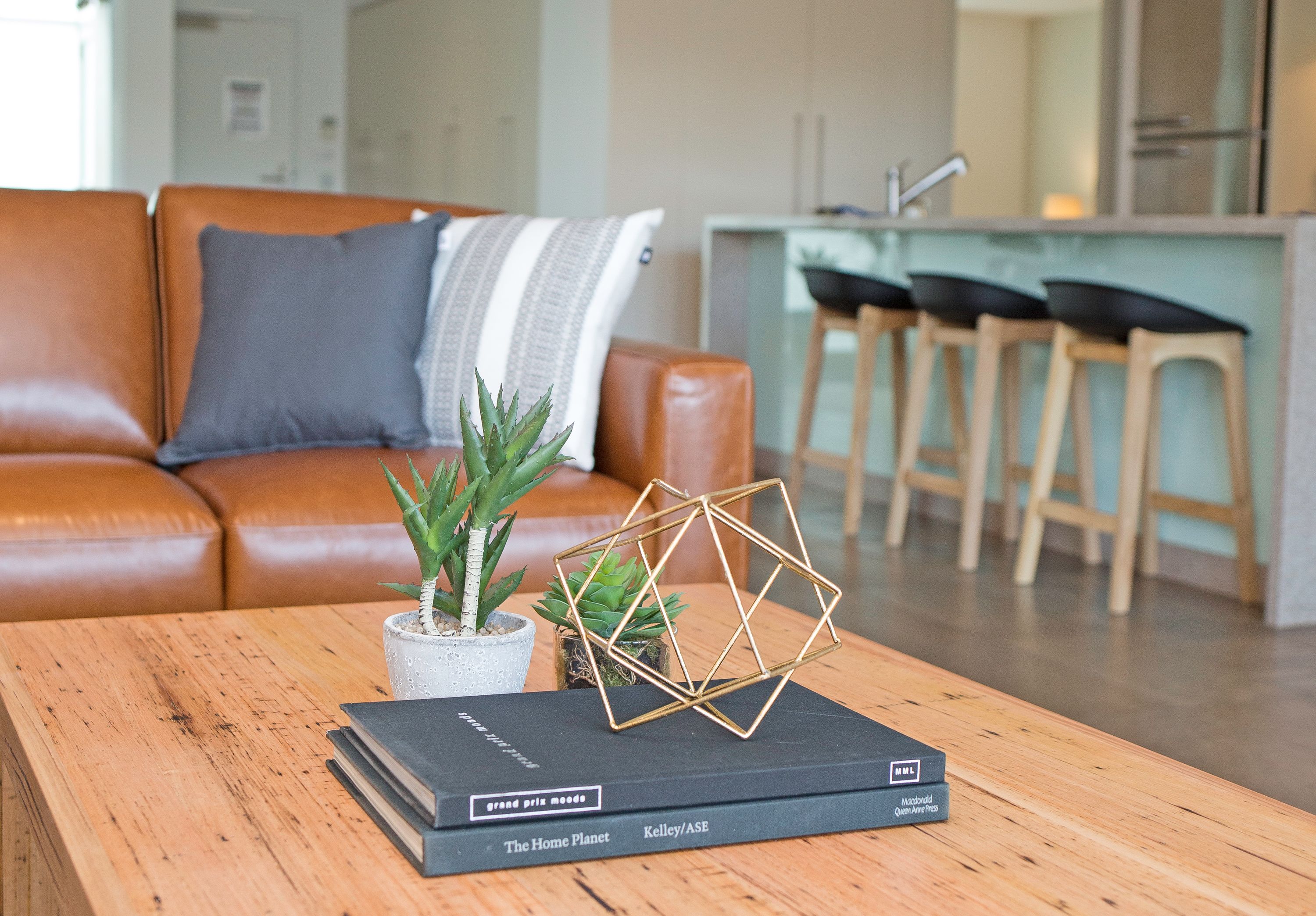 Een moderne woonkamer met een leren bank, een bijzettafeltje met boeken en een geometrisch kunstwerk, en een keuken op de achtergrond.