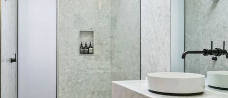 A modern bathroom with a round sink, a handheld showerhead, and a built-in shelf with toiletries.