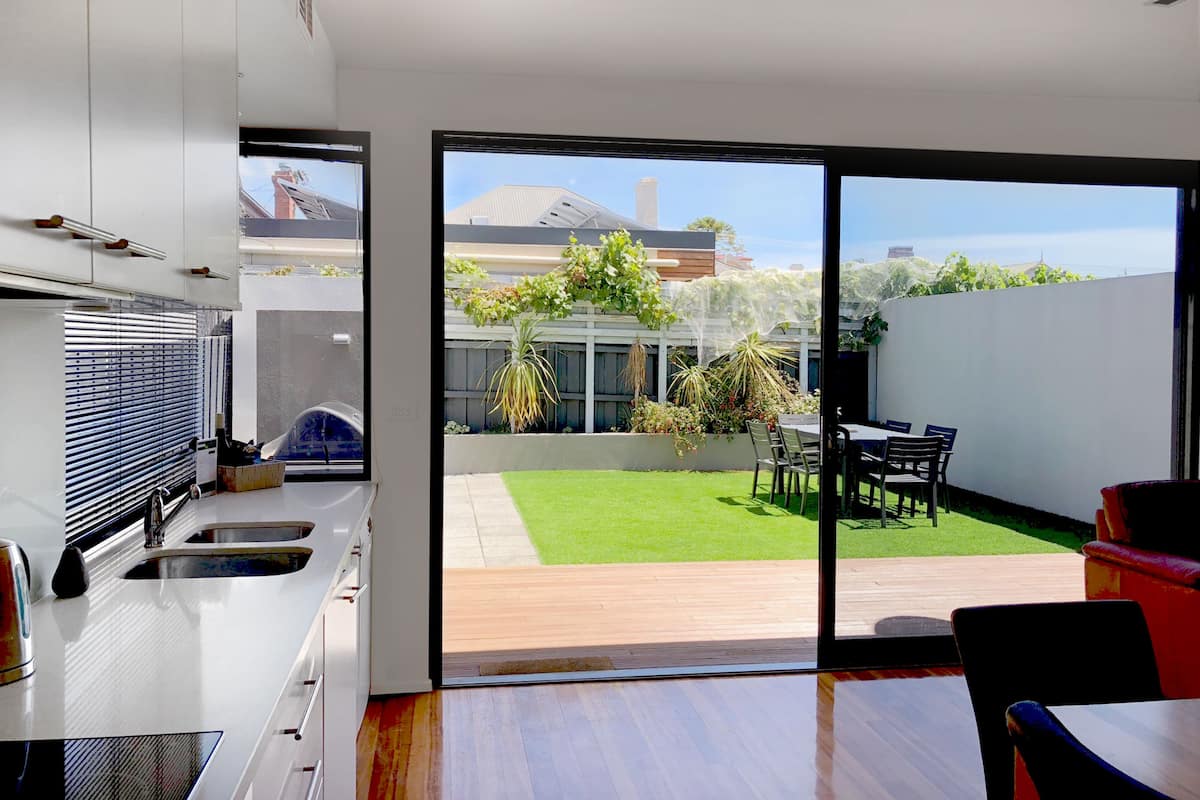A modern kitchen with a large sliding glass door leading to a backyard with a wooden deck, a dining area, and a small garden.