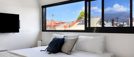 A bedroom with a large window, a bed with white bedding, and a TV mounted on the wall.