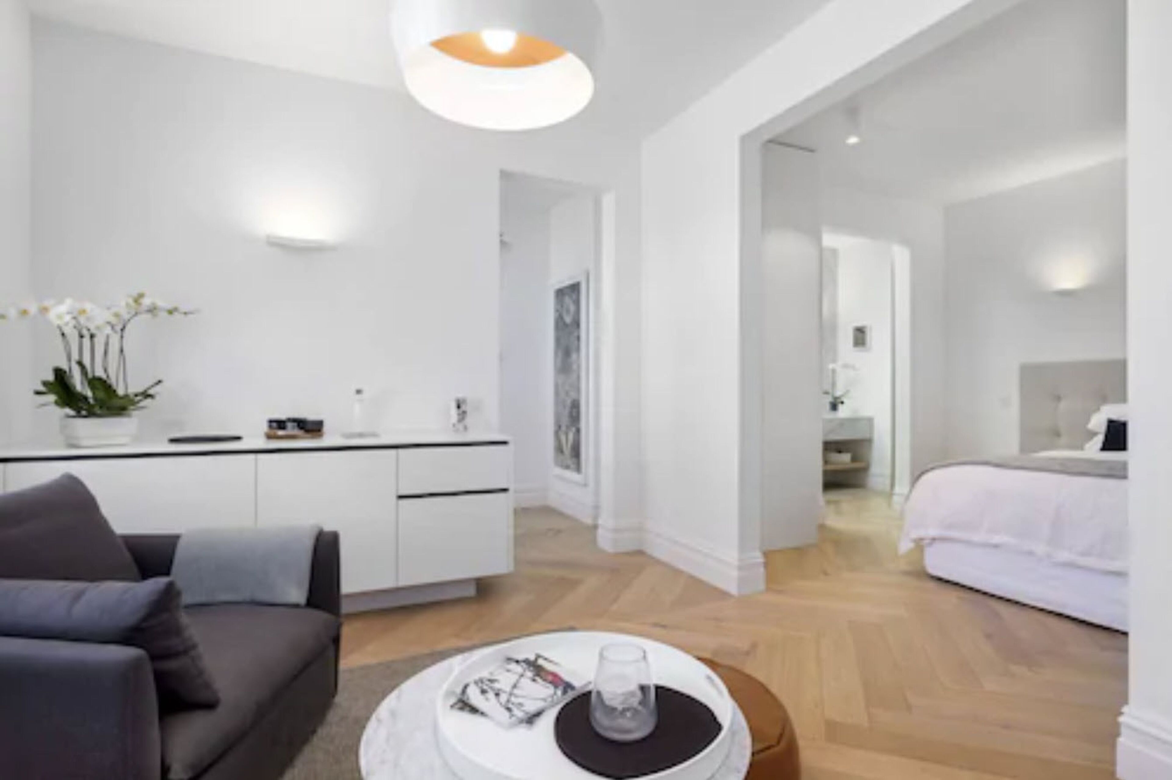 Une chambre d’hôtel moderne et minimaliste, avec un canapé gris, une petite table ronde en verre et un lit recouvert de linge de lit blanc.