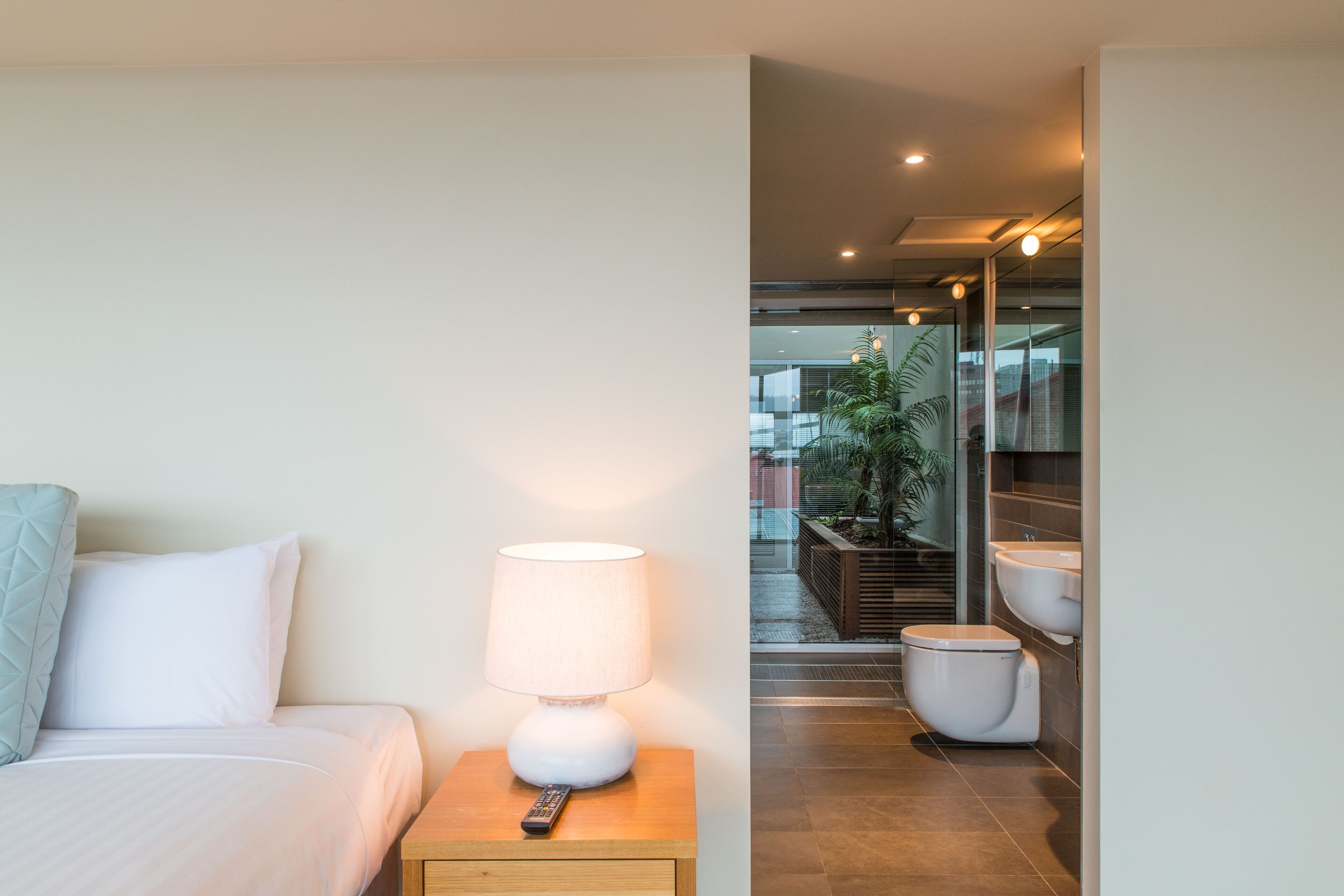 A hotel room with a bed, bedside lamp, and a remote control on a wooden nightstand.
