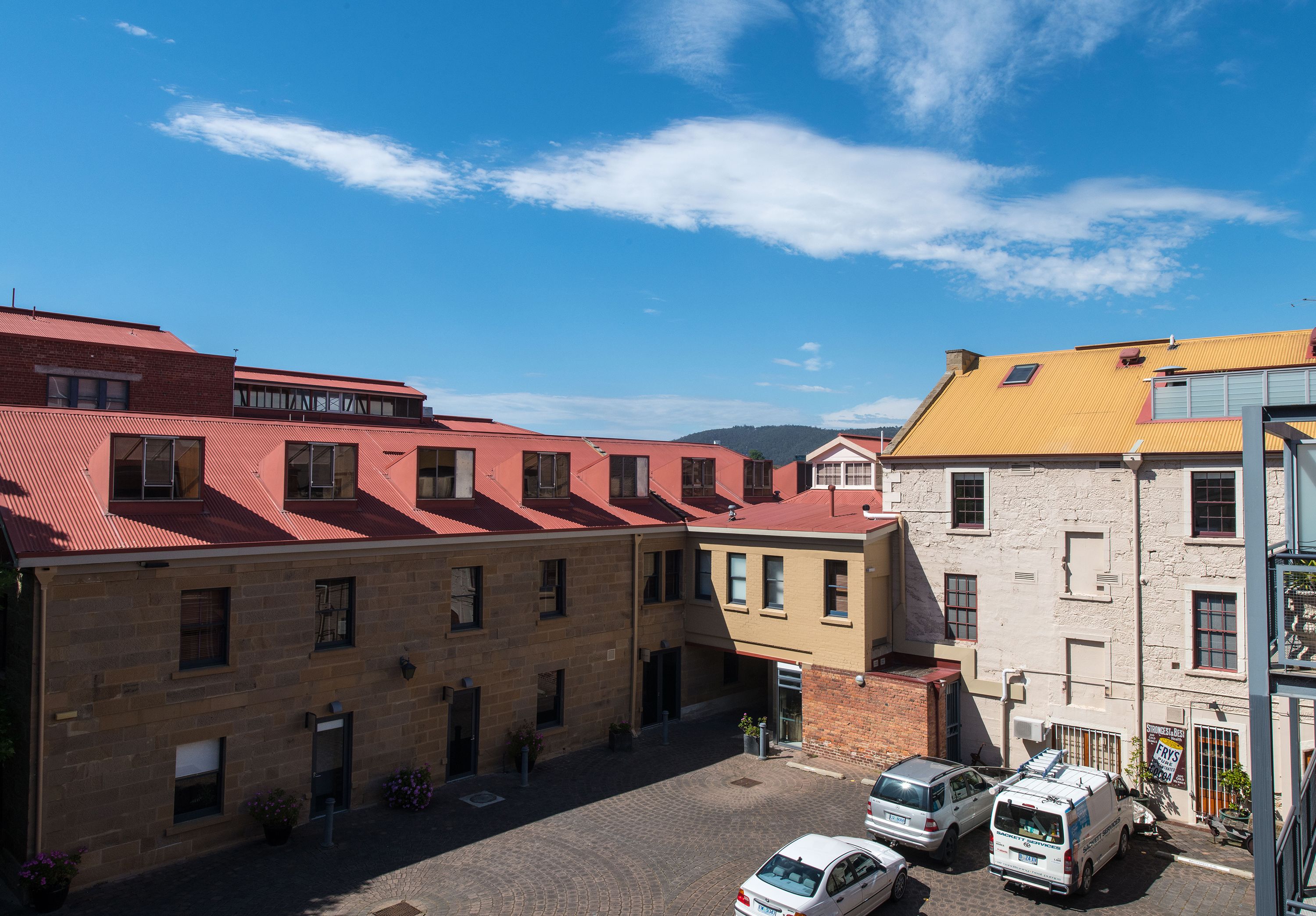 Un immeuble à plusieurs étages avec un toit rouge, un bâtiment jaune attenant, et une cour pavée où sont garées des voitures et un fourgon.