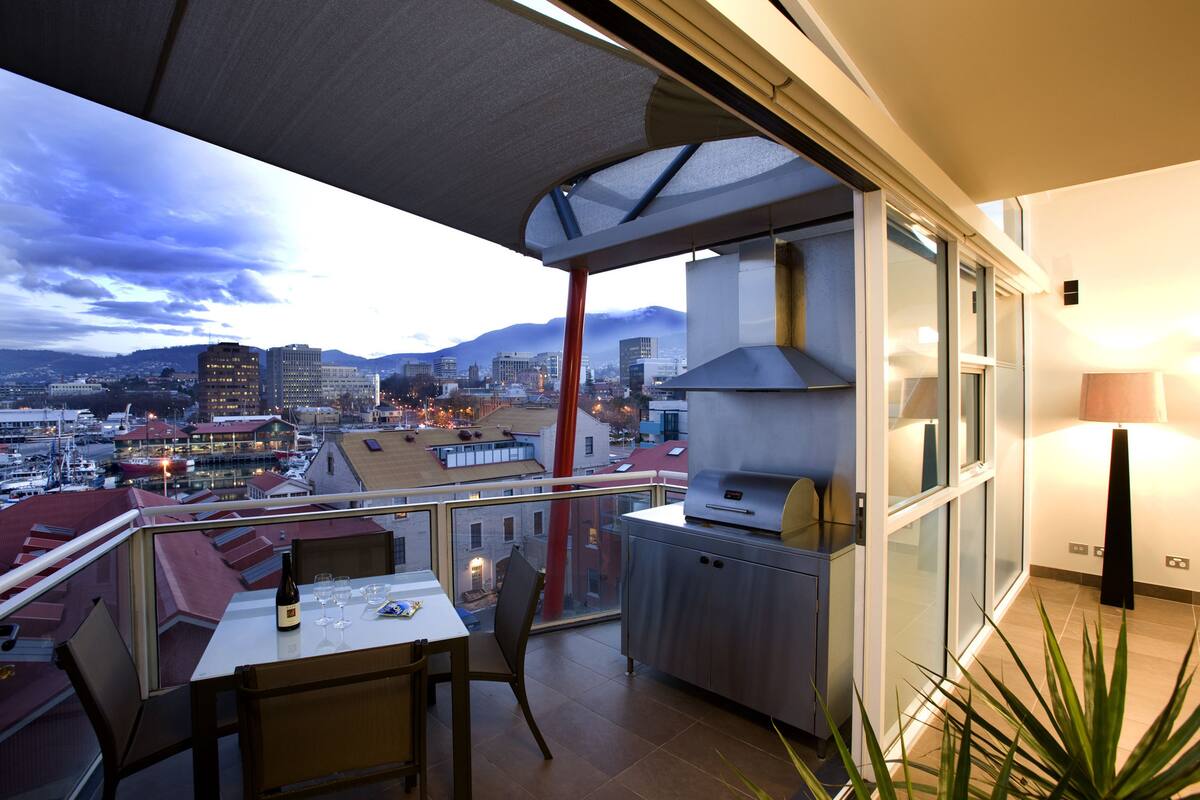 A balcony with a table and chairs, a grill, and a view of the cityscape and mountains.