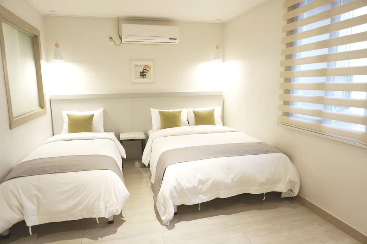 A hotel room with two beds, a nightstand, an air conditioning unit, a window with blinds, and a framed picture on the wall.