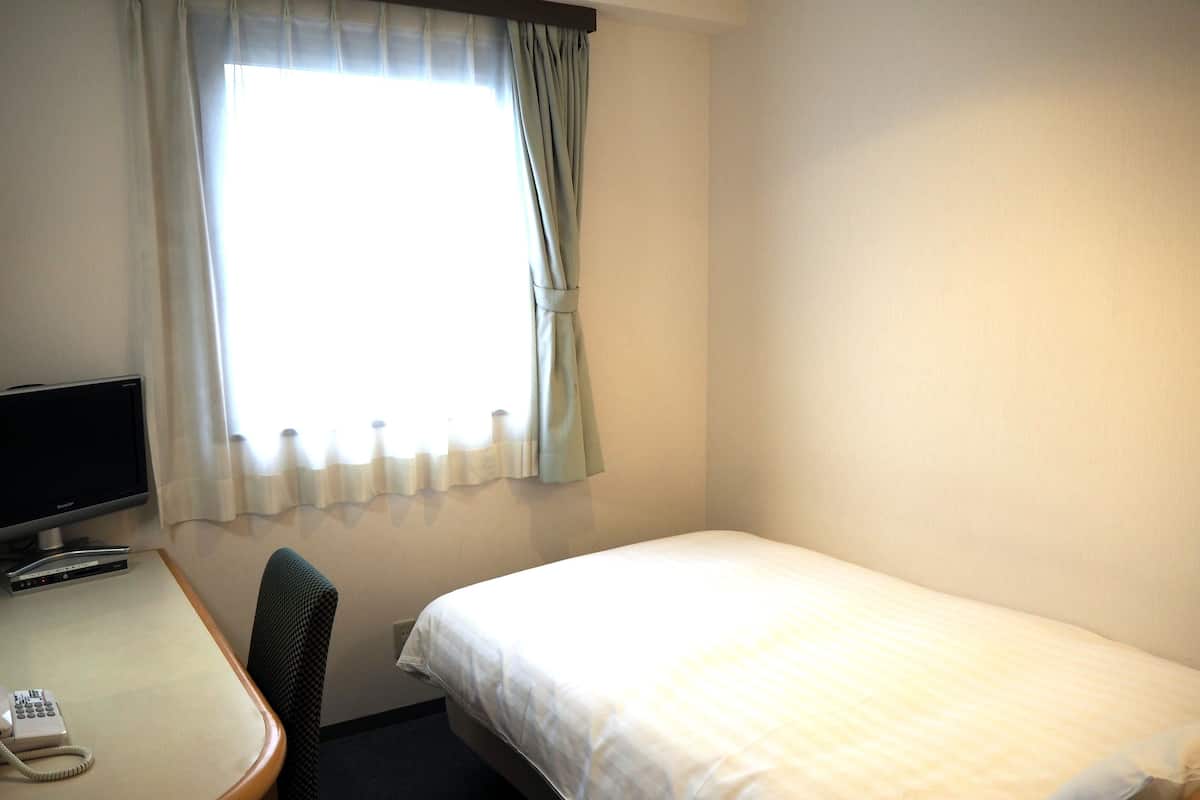 Quarto de hotel com uma cama, uma escrivaninha com computador e telefone, e uma janela com cortinas.