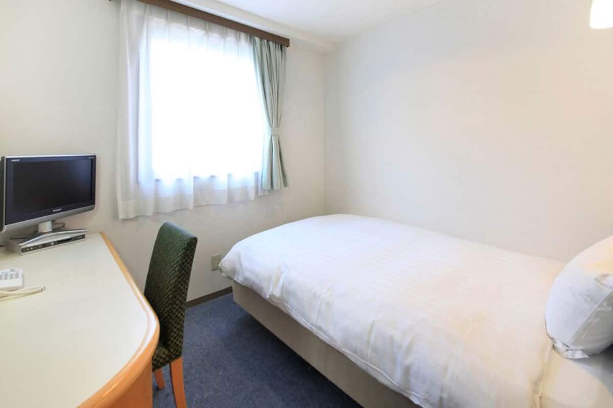 Quarto de hotel com cama, escrivaninha com computador, cadeira e janela com cortinas.