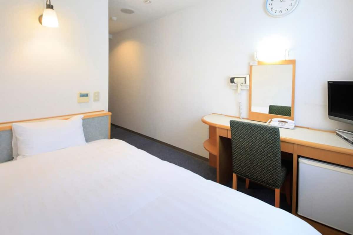 Quarto de hotel com cama, escrivaninha, cadeira, televisão, espelho e relógio na parede.