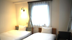 Ein Hotelzimmer mit zwei Einzelbetten, einem kleinen runden Tisch, einem Stuhl, einer Lampe und einem Fenster mit Vorhängen.