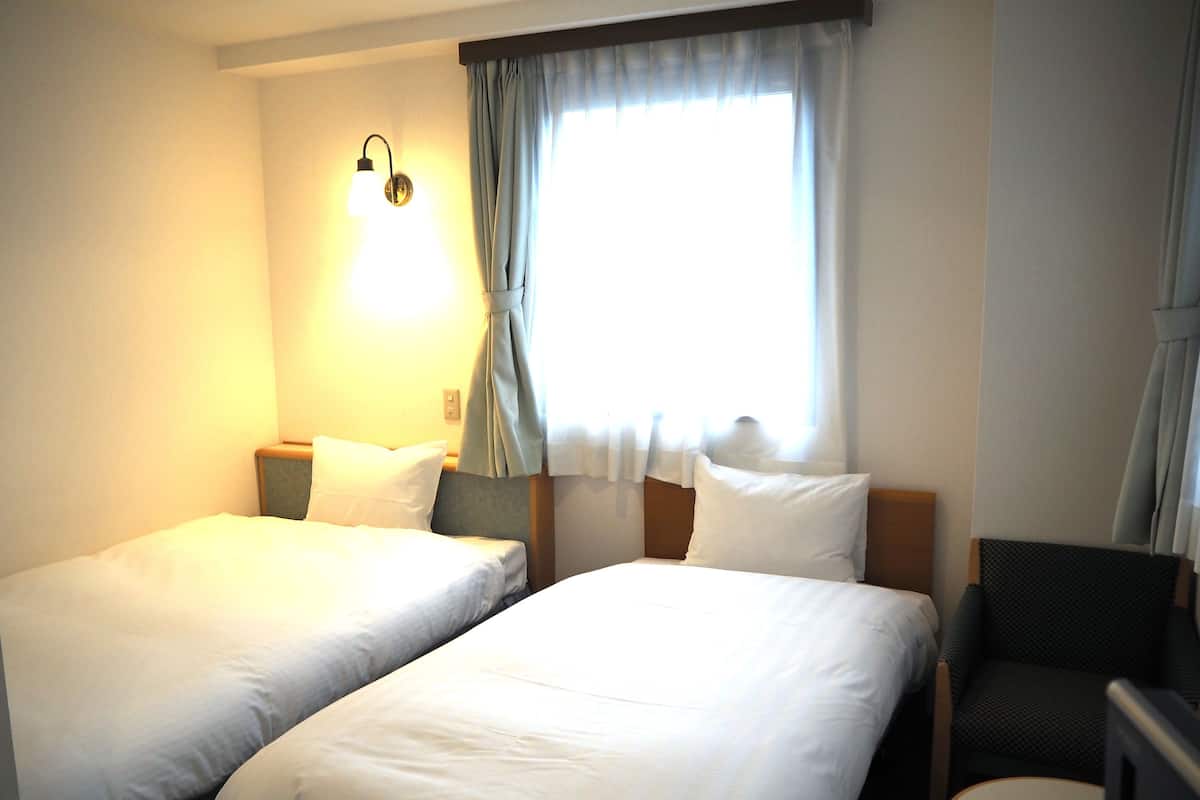 Quarto de hotel com duas camas de solteiro, uma mesa redonda pequena, uma cadeira, um abajur e uma janela com cortinas.