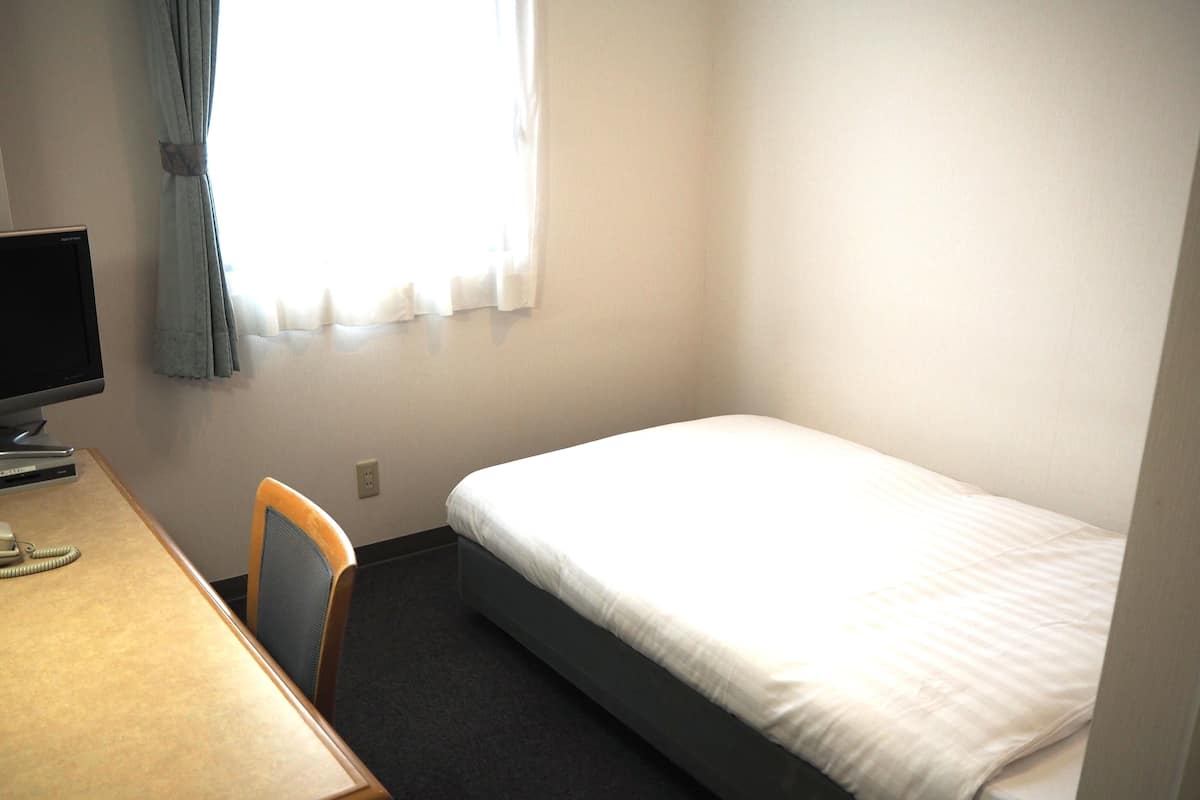 Quarto pequeno e limpo com uma cama de solteiro, uma escrivaninha com computador e uma cadeira.