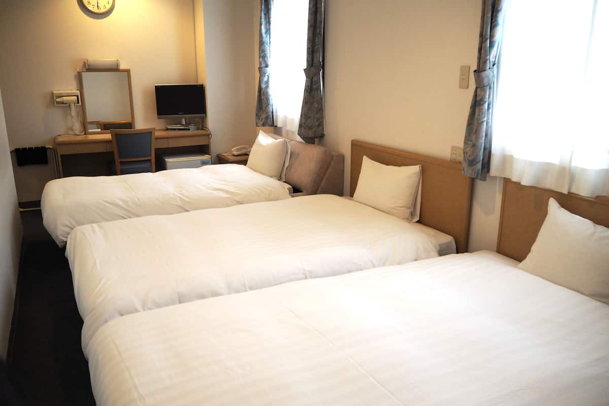 Quarto de hotel com três camas de solteiro, uma mesa de trabalho com computador e um relógio na parede.