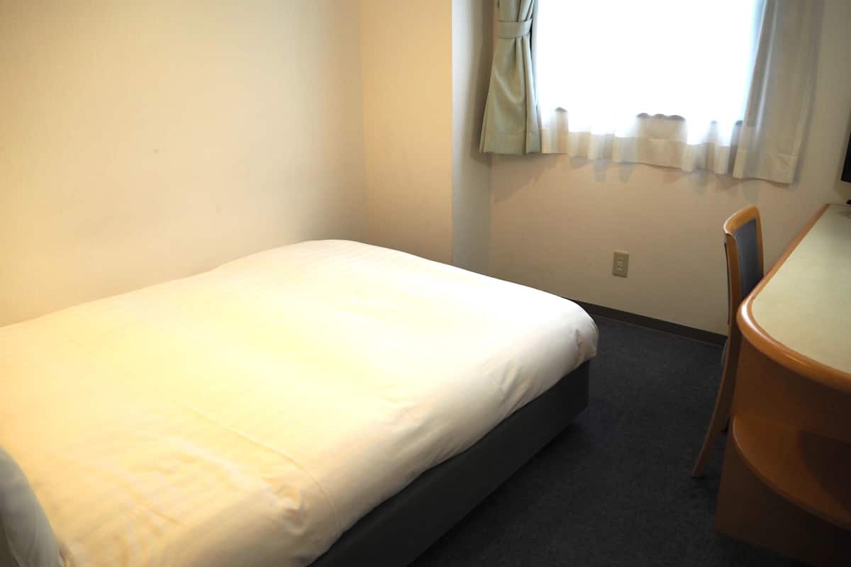 Cama de solteiro com colchão branco e cabeceira escura em quarto pequeno com janela coberta por cortinas leves.