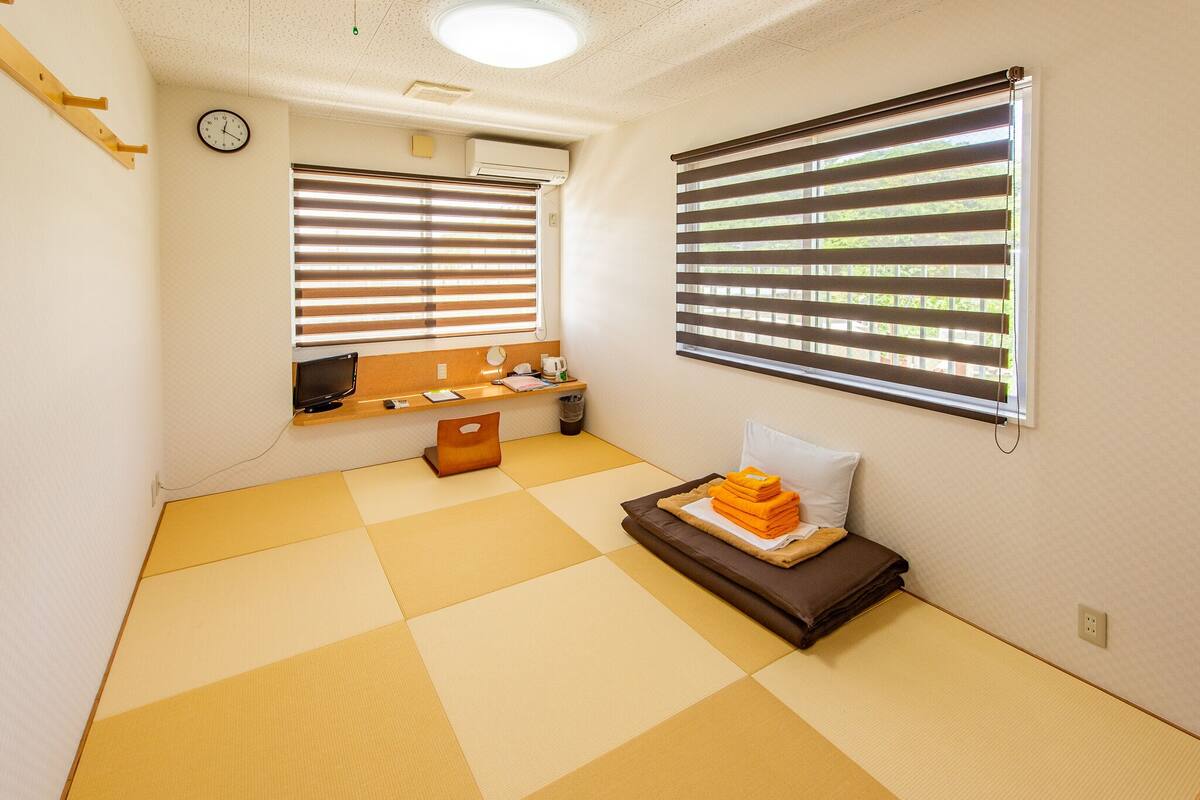 Een kamer met tatami matten, twee schuiframen, een klok en een klein plankje met een waterkoker en een kop.