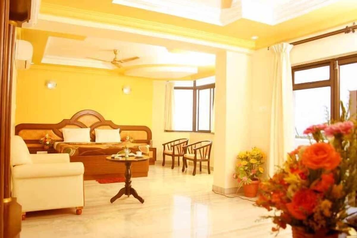 A hotel room with a bed, a sofa, a dining table, and chairs. There is a window with curtains and a ceiling fan.
