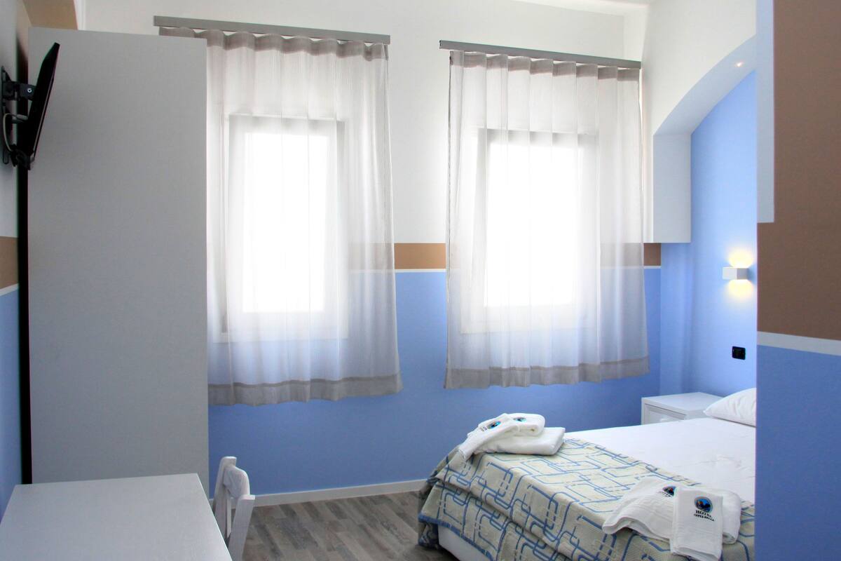 Habitación de hotel con dos ventanas, una cama con colcha estampada, una mesita de noche y una silla.