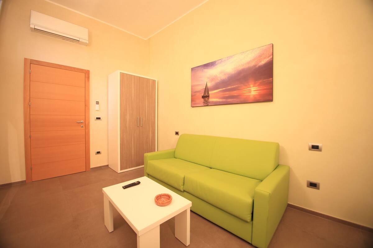 Una habitación con un sofá verde, una mesa blanca, un cuadro de un atardecer y una puerta de madera.