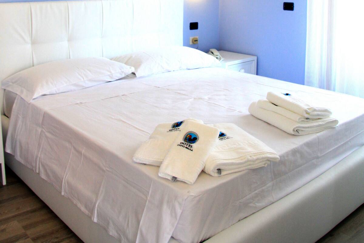 Una habitación de hotel con una cama grande, ropa de cama blanca y una pared azul.