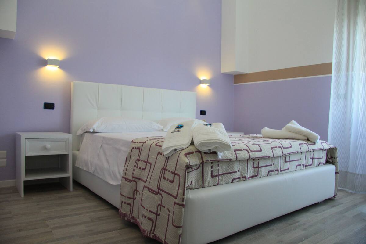 Una cama bien hecha con un edredón estampado, una mesita de noche blanca y dos lámparas fijadas a la pared.