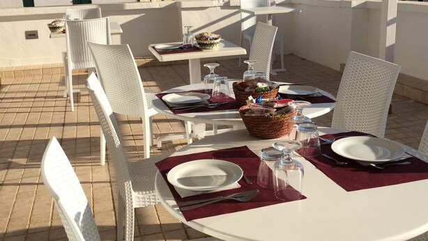 A covered outdoor dining area with white wicker furniture, a table set with plates, cutlery, and a basket of bread. - Hotel Città Bella (Gallipoli)